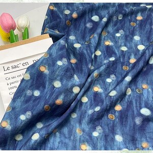 May include: A blue fabric with a pattern of white and gold circular designs. The fabric is draped, showing folds and texture. The text "Le sac en pa" is visible on a paper bag.