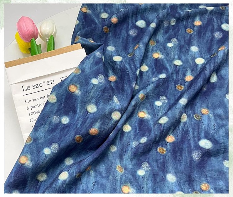 May include: A blue fabric with a pattern of white and gold circular designs. The fabric is draped, showing folds and texture. The text "Le sac en pa" is visible on a paper bag.