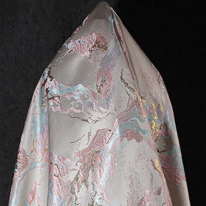 May include: A close-up of a light gray fabric with a pink, blue, and gold marble pattern. The fabric has a subtle shimmer and a soft, luxurious feel.