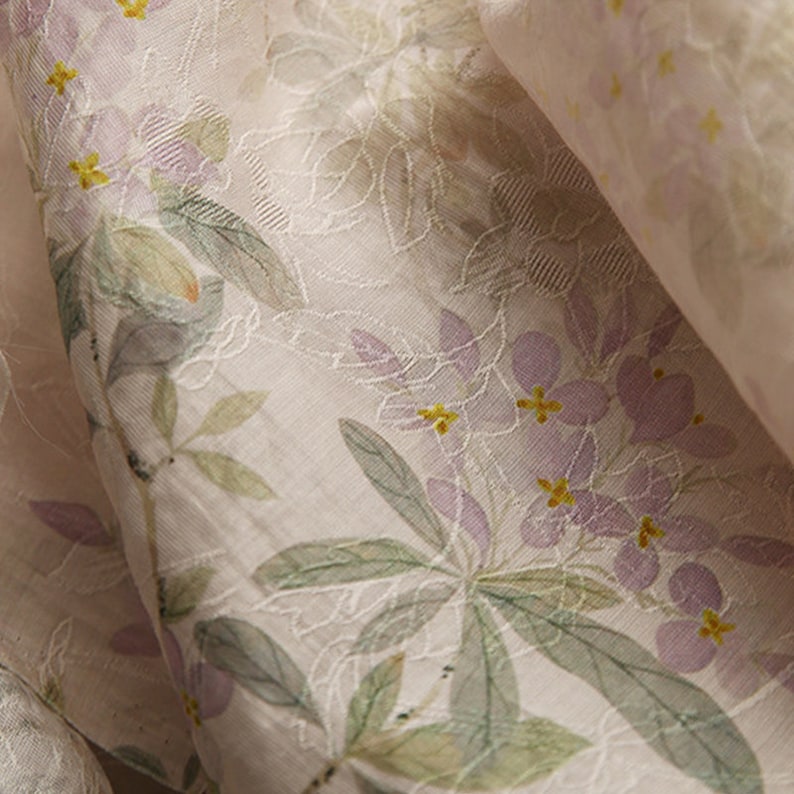 Pu&ograve; includere: Primo piano di un tessuto trasparente con stampa floreale. Il tessuto &egrave; di colore beige chiaro con una texture sottile. I fiori sono di un viola pallido con centri gialli. Le foglie sono di un verde chiaro.
