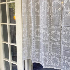 May include: A sheer light gray curtain panel featuring a repeating grid pattern of white embroidered floral wreaths. The fabric is constructed from joined square sections separated by delicate openwork lace detailing throughout the design.