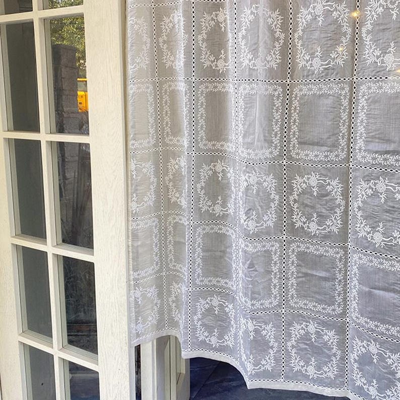 May include: A sheer light gray curtain panel featuring a repeating grid pattern of white embroidered floral wreaths. The fabric is constructed from joined square sections separated by delicate openwork lace detailing throughout the design.