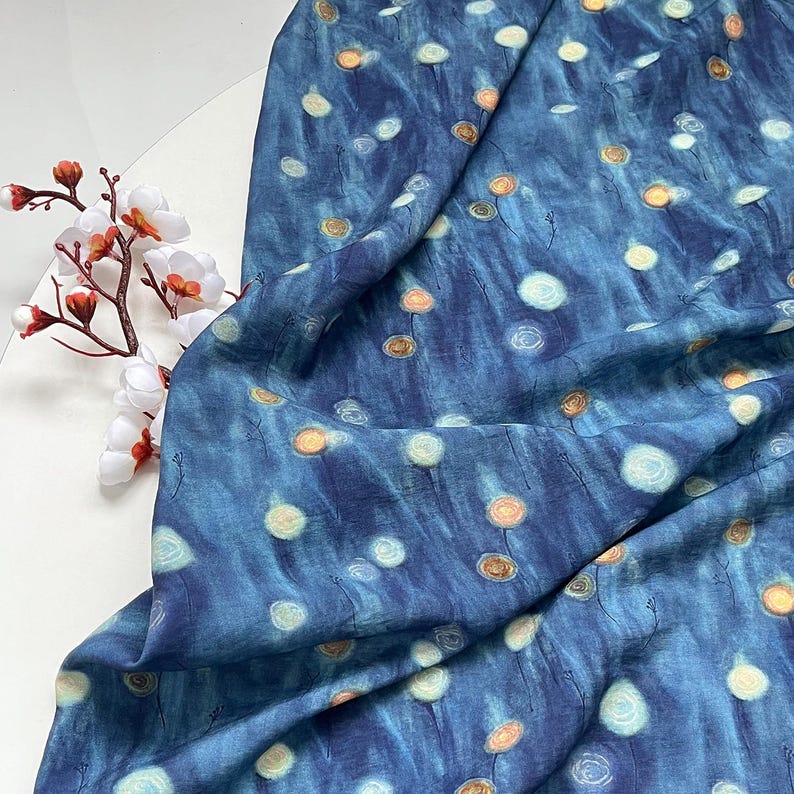 May include: A close-up of a blue fabric with a pattern of white and orange floral designs. The fabric is draped, showing the texture and flow of the material. The floral pattern is scattered across the fabric.