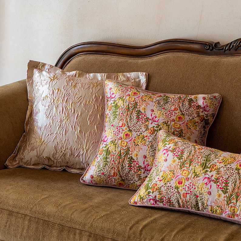 May include: Three decorative throw pillows on a brown sofa. The pillow at the back is a light pink with a textured pattern. The two pillows at the front are pink with a floral pattern.