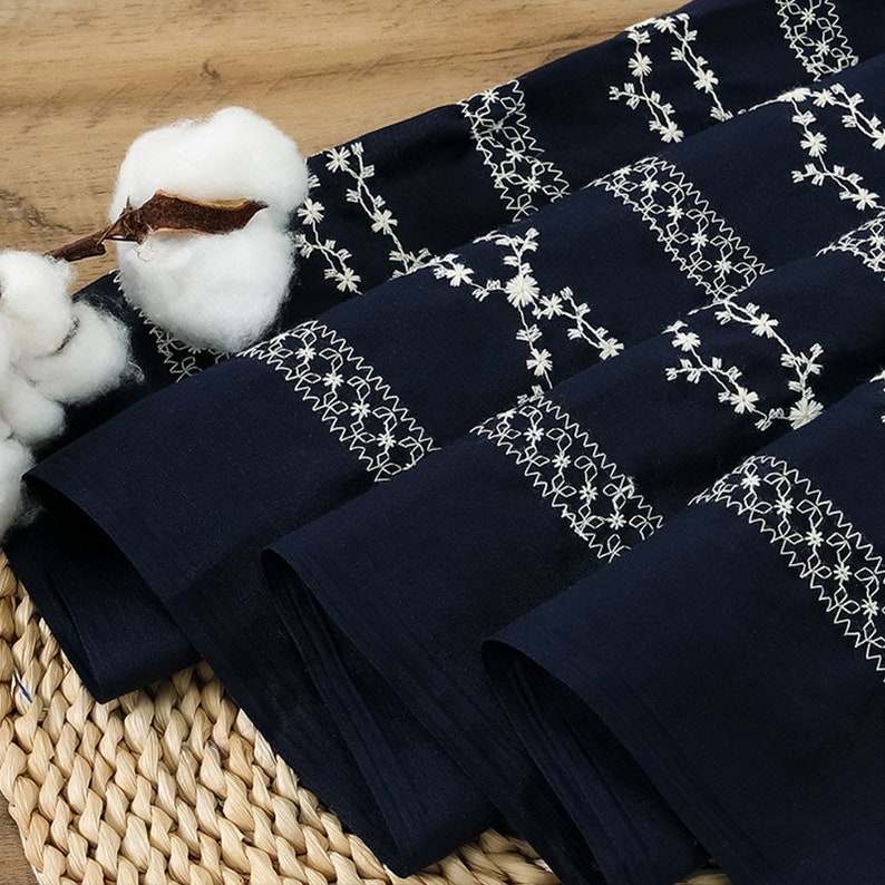 May include: Close-up of navy blue fabric with white embroidered floral patterns. The fabric is folded and stacked, showing the intricate details of the embroidery.