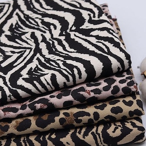 May include: A stack of four fabric swatches featuring different animal print patterns. The top swatch is a black and white zebra print, the second is a pink and brown leopard print, the third is a brown and black leopard print, and the bottom swatch is a beige and black zebra print.