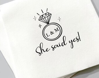 May include: White cocktail napkins with a black diamond ring graphic and the text "She said yes!". The ring design includes the initials "L & M" inside a circle. Ideal for engagement parties and weddings.