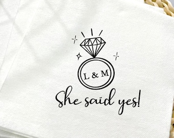 May include: White paper napkins with a black diamond ring graphic and the text "She said yes!" The ring design includes the initials "L & M" inside a circle. Napkins are stacked on a woven surface.