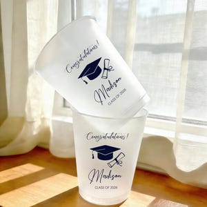 May include: Two white plastic cups stacked on a wooden surface. Each cup has the text "Congratulations!" above a graduation cap and diploma graphic, with the name "Madison" and "CLASS OF 2026" printed below in navy blue.