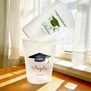 Può includere: Due bicchieri di laurea in plastica bianca. Una tazza ha il nome "Madison" e le parole "Congratulazioni!" e "Classe 2026" stampate in verde. L'altra tazza ha il nome "Angela" e "Classe 2026" stampate in nero.