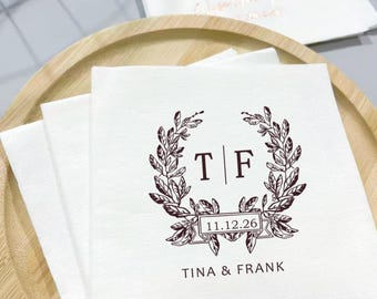 May include: White paper napkins featuring a brown floral wreath design, the initials "T | F", and the date "11.12.26". The names "TINA & FRANK" are printed below. The napkins are on a wooden tray.
