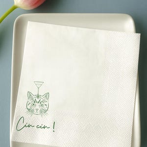 May include: White cocktail napkin featuring a green illustration of a cat with a martini glass. The napkin has the phrase "Cin cin!" in green script. It rests on a white rectangular tray, with a pink tulip in the top left corner.