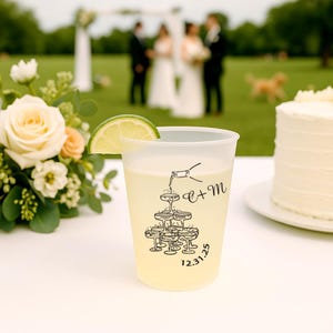 Può includere: Un bicchiere di plastica trasparente pieno di una bevanda gialla e una fetta di lime. La tazza presenta una grafica nera di calici di champagne e una bottiglia che versa, con le iniziali "E + M" e la data "12.31.25". Una torta nuziale e fiori sono sullo sfondo.