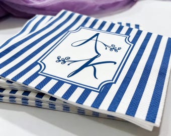 May include: A stack of blue and white striped paper napkins. Each napkin has a white rectangular emblem with the cursive letters "A" and "K" in blue, with floral accents. The napkins are stacked on a white surface.