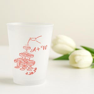 May include: A frosted plastic cup featuring a red illustration of champagne glasses arranged in a pyramid, with a bottle pouring champagne. The cup has the initials "A + W" and the date "2.28.26" printed in red.