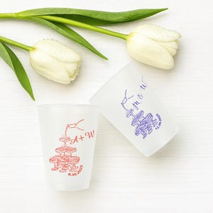 Può includere: Due bicchieri di plastica smerigliata con illustrazioni a tema matrimonio. Una tazza presenta un'illustrazione rossa di champagne versato in una torre di bicchieri, con le iniziali "A + W" e la data "8.30.2025". L'altra tazza ha un'illustrazione blu con le iniziali "M & W" e la data "12.30.2025".