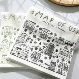 May include: A stack of white paper napkins with a black and gray illustrated map design. The napkins are printed with the text "MAP OF US" and the names "MICHELLE & DANIEL". The map includes illustrations of buildings, trees, and landmarks.