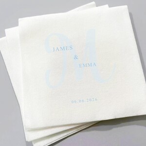 May include: A stack of white paper napkins with light blue text. The text includes the names "JAMES & EMMA" and the date "06.06.2026". The napkins are folded and stacked neatly.