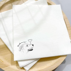 May include: White paper napkins with a black line drawing of a dog balancing a wine glass on its nose. The text "Drinks on me!" is written in a script font. The napkins are stacked on a wooden plate.