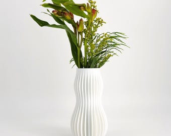 May include: A white vase with a vertical ribbed texture and a curved, hourglass-like silhouette. The vase contains a bouquet of green foliage, including long pointed leaves, small yellow-green buds, and thin, feathery stems arranged upright.
