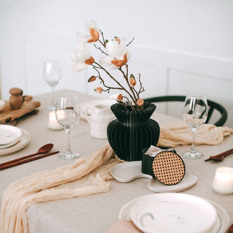 K&ouml;nnte beinhalten: Eine schwarze Vase im Akkordeon-Stil mit wei&szlig;en Magnolienbl&uuml;ten auf einem Tisch mit Weingl&auml;sern, wei&szlig;en Tellern, Holzl&ouml;ffeln, einem beigen Tischl&auml;ufer und einem kleinen schwarzen Beh&auml;lter mit einem runden Rattandeckel auf einem wei&szlig;en Tablett.