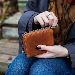 May include: A brown leather zipped wallet with a gold zipper. The wallet is being held in someone's hand.