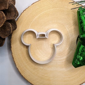 May include: A silver metal cookie cutter shaped like Mickey Mouse's head. The cutter is sitting on a wooden surface.