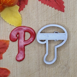 May include: A red plastic letter "P" and a silver cookie cutter in the shape of a baseball cap. The cookie cutter has a raised line across the top of the cap.