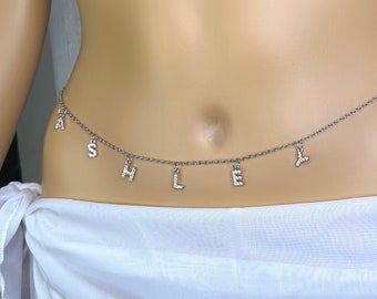 May include: A silver-toned belly chain with the name "ASHLEY" spelled out in individual letter charms. The letters are embellished with small, sparkling accents. The chain is draped across the lower abdomen, creating a delicate and personalized accessory.