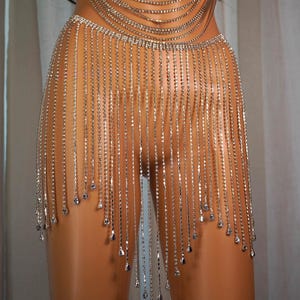 以下が含まれることがあります： シルバーのラインストーンボディチェーンとスカートのセット。上部は肩と胸にドレープし、スカートはウエストから垂れ下がっています。デザインは複数のラインストーンのストランドを特徴とし、端にはより大きな涙型のラインストーンが付いています。