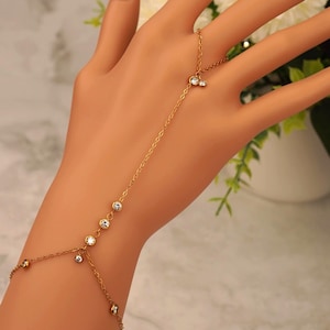 May include: A gold-colored hand chain bracelet with clear, round gemstones. The bracelet connects to a ring and features multiple gemstones along the chain. The hand is positioned in front of a blurred background of white flowers and greenery.