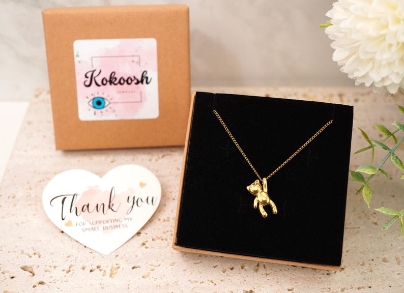 May include: A gold bear pendant necklace presented in a black jewellery box. The necklace hangs on a gold chain. A brown box with a sticker reading "Kokoosh" is in the background. A heart-shaped card with "Thank you" is also visible.