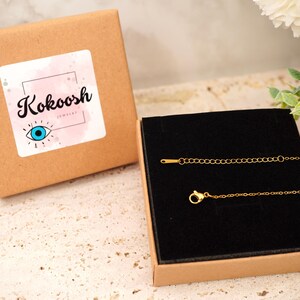 May include: A gold necklace with a fine chain and clasp, displayed in a black velvet-lined box. The box is inside a brown cardboard box with a sticker reading "Kokoosh Jewelry" and an eye symbol.