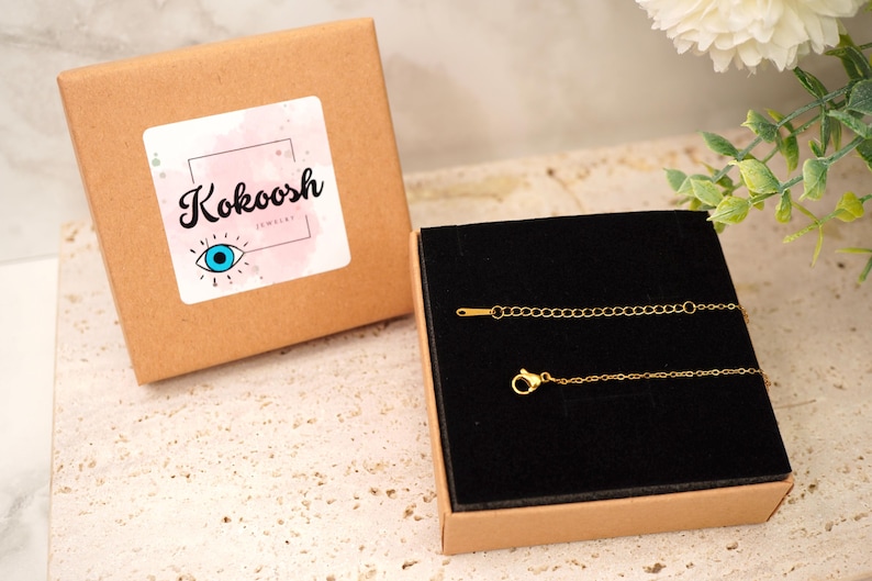 May include: A gold necklace with a fine chain and clasp, displayed in a black velvet-lined box. The box is inside a brown cardboard box with a sticker reading "Kokoosh Jewelry" and an eye symbol.