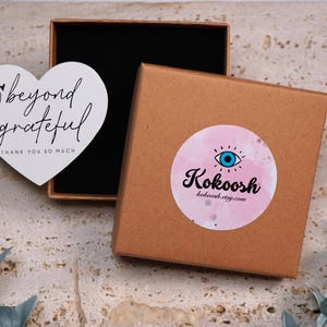 May include: A brown cardboard box with a black interior, next to a white heart-shaped card with the words "beyond grateful" and "thank you so much". A round sticker with a pink watercolor background and an eye design is on the box.