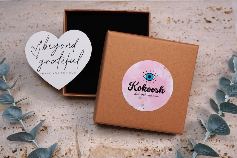 May include: A brown cardboard box with a black interior, next to a white heart-shaped card with the words "beyond grateful" and "thank you so much". A round sticker with a pink watercolor background and an eye design is on the box.