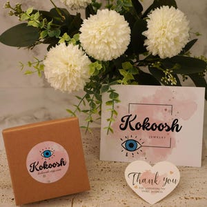 May include: A jewellery box with a pink and blue logo, a card with the brand name "Kokoosh Jewelry", and a heart-shaped thank you note. The scene is set against white flowers and greenery, creating a soft, inviting backdrop.