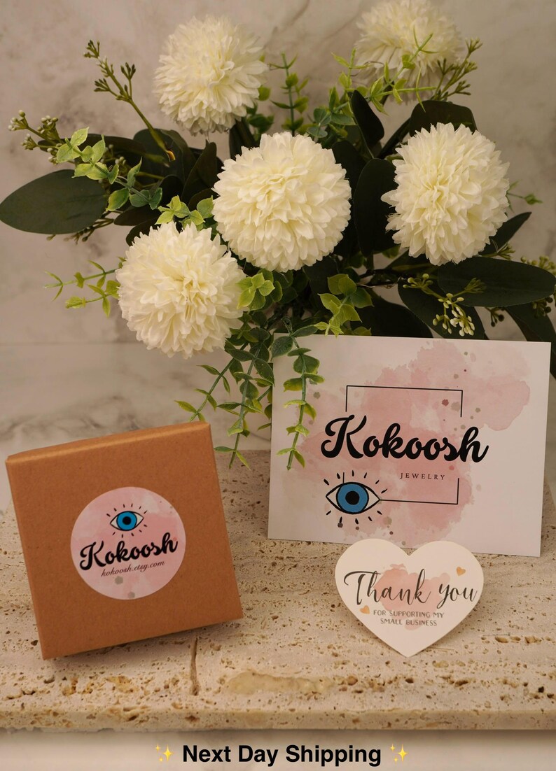 May include: A jewellery box with a pink and blue logo, a card with the brand name "Kokoosh Jewelry", and a heart-shaped thank you note. The scene is set against white flowers and greenery, creating a soft, inviting backdrop.