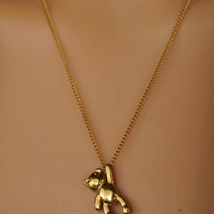 May include: A gold-coloured necklace with a small teddy bear pendant. The bear is suspended from the chain, with its arms raised. The chain is a delicate, gold-toned curb chain. The background is a neutral, skin-toned colour.