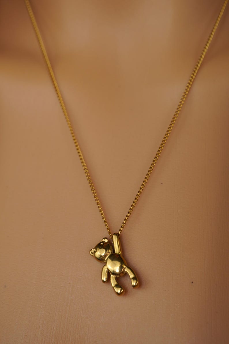 May include: A gold-coloured necklace with a small teddy bear pendant. The bear is suspended from the chain, with its arms raised. The chain is a delicate, gold-toned curb chain. The background is a neutral, skin-toned colour.