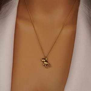 May include: A gold-coloured necklace with a small teddy bear pendant. The pendant hangs from a delicate chain. The necklace is worn against a white blazer.