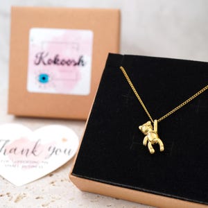 May include: A gold-coloured necklace with a small bear pendant suspended from a fine chain. The necklace is presented in a black box, with a brown box and a heart-shaped "Thank you" card in the background. The brand name "Kokoosh" is visible.