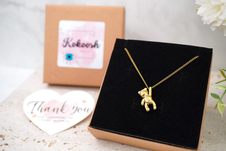 May include: A gold-coloured necklace with a small bear pendant suspended from a fine chain. The necklace is presented in a black box, with a brown box and a heart-shaped "Thank you" card in the background. The brand name "Kokoosh" is visible.