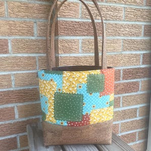 May include: A patchwork tote bag with a cork base and handles. The bag features a colorful floral pattern with yellow, blue, and green squares. The handles are a light brown color, and the bag is sitting on a wooden surface.