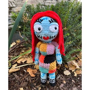 Op de afbeelding: Een gehaakte pop met rood garen haar, een blauw-witte patchwork jurk en zwart-witte geruite schoenen. De pop staat op een bed van bruine houtsnippers.