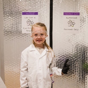 May include: A young girl wearing a white lab coat, safety goggles, and black gloves stands in front of a door with a sign that reads "SECRET LAB Authorized Personal Only".