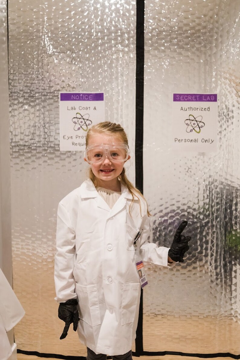 May include: A young girl wearing a white lab coat, safety goggles, and black gloves stands in front of a door with a sign that reads "SECRET LAB Authorized Personal Only".