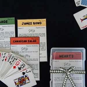 May include: A collection of card game instructions and playing cards on a black surface. The instructions are for games like "Garbage", "James Bond", "Canadian Salad", and "Hearts". A deck of cards and a clear plastic case with a green and white ribbon are also present.
