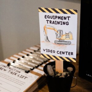 May include: A yellow and black sign with the text "Equipment Training" and a cartoon excavator. The sign is propped up in a small black bucket filled with small rocks. The text "Video Center" is below the excavator.