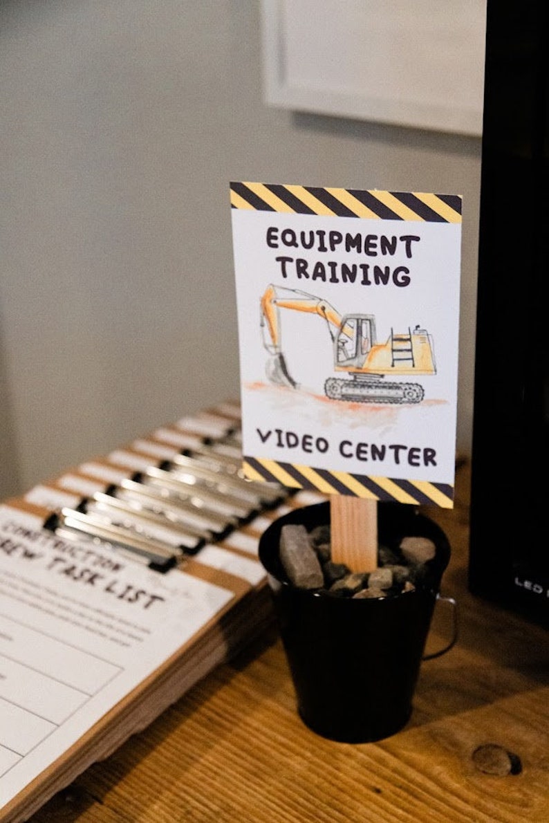 May include: A yellow and black sign with the text "Equipment Training" and a cartoon excavator. The sign is propped up in a small black bucket filled with small rocks. The text "Video Center" is below the excavator.